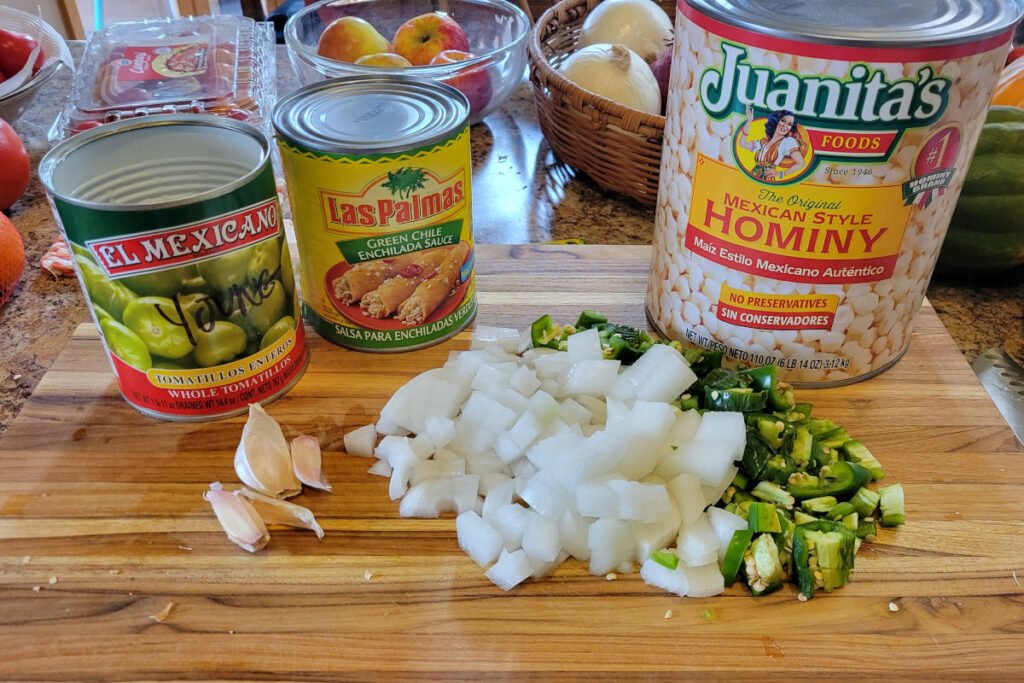 Ingredients for chicken pozole verde — tomatillos, green chile sauce, hominy, garlic, onions, and jalapeños on a wooden cutting board.