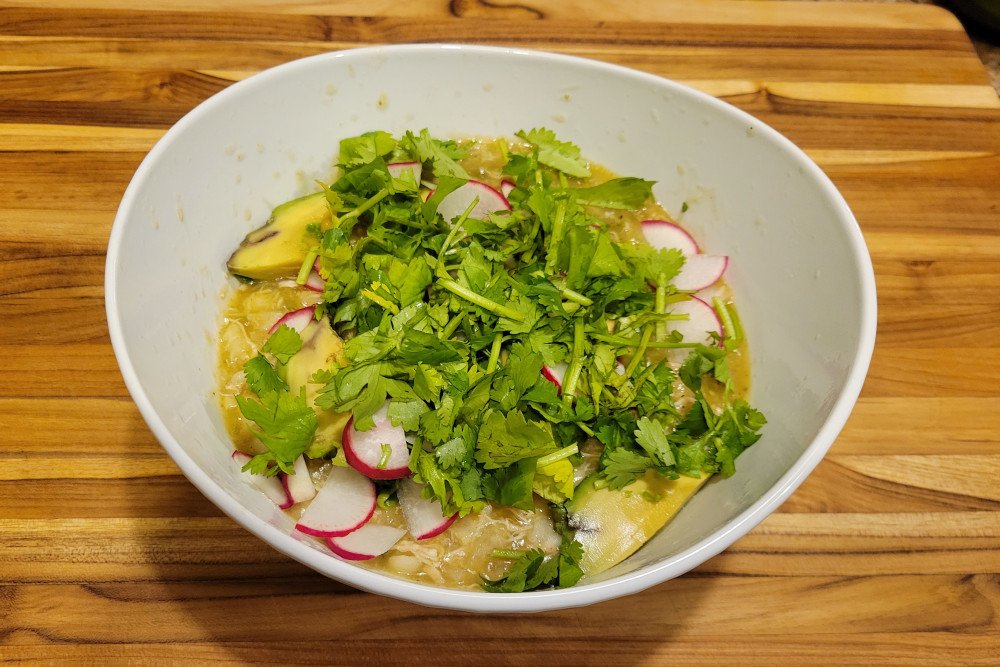 Bowl of chicken pozole verde topped with cilantro, radishes, and avocado slices.
