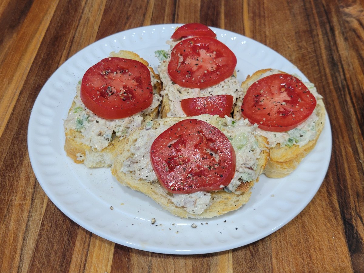 Turkey salad crostini on a white plate, topped with fresh tomato slices and cracked black pepper.