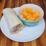 Turkey salad wrap sliced and served on a white plate with a bowl of fresh cantaloupe.