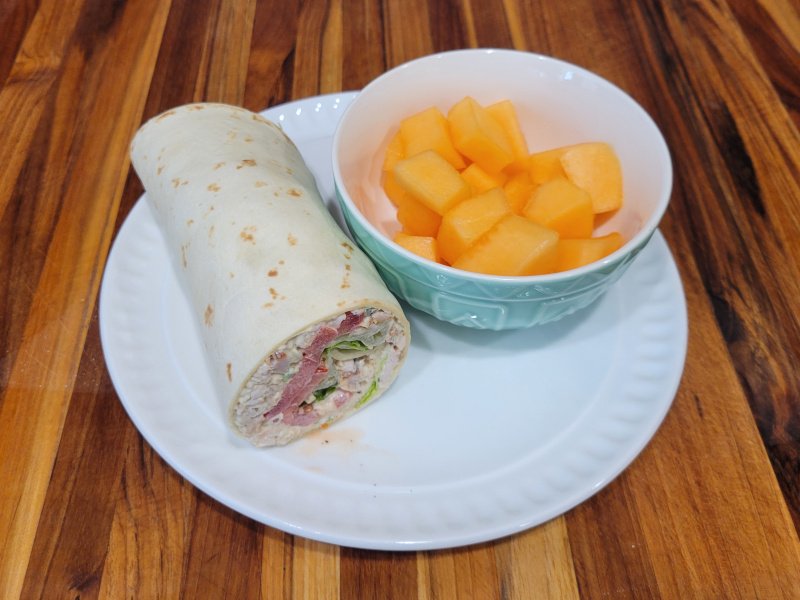 Turkey Salad Wrap – Step 5 Plated Turkey salad wrap sliced and served on a white plate with a bowl of fresh cantaloupe.
